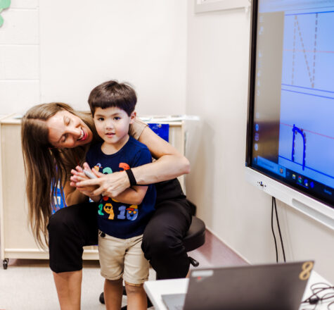 Boy sitting on instructor therapists lap in front of a LED screen with school work on it
