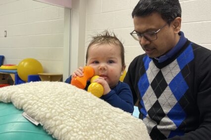 baby boy stands with the support of a therapy ball and holds toys close to his face. He is supported to stand by a male therapist.