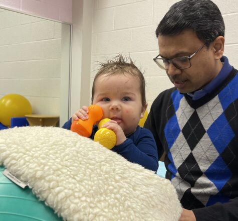 baby boy stands with the support of a therapy ball and holds toys close to his face. He is supported to stand by a male therapist.